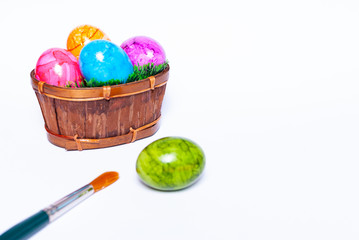 A basket filled with green grass and colorful easter eggs, on egg fallen out and a paintbrush on white background with copy space