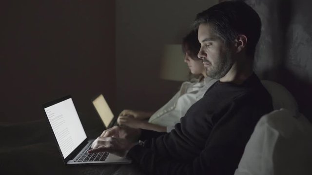 Couple typing on laptops in bedroom at night. Side view of focused young man and woman using laptops together in bed. Technology concept