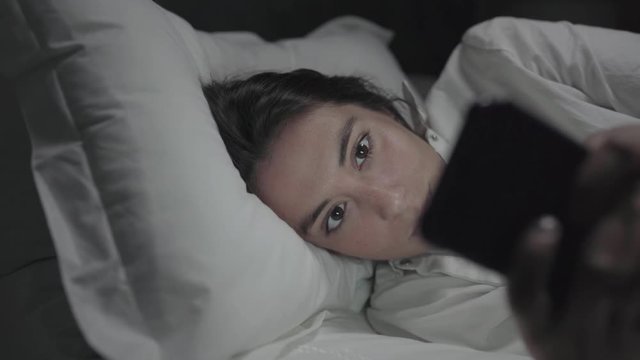 Tired young woman using cell phone in bed. Beautiful sleepy girl lying in bed and using mobile phone at night. Connection concept
