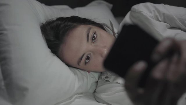Sleepy young woman using smartphone in bed. Beautiful smiling girl lying in bed and using cell phone at night. Communication concept