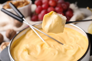 Piece of bread over pot with delicious cheese fondue on table