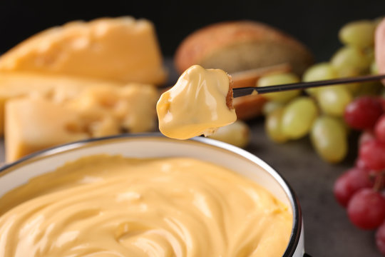 Piece of bread over pot with delicious cheese fondue, closeup