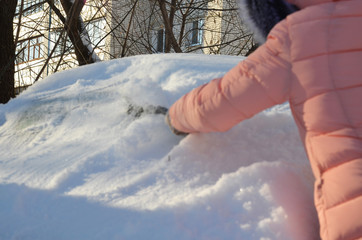 Winter portrait of a young woman cleaning snow from a car. Beauty blonde Model Girl laughs and cheerfully cleans the snow. Beautiful young woman outdoors.