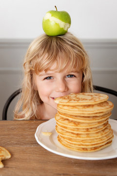 Smiling little girl hiding behing stack of pancakes with an apple on top of her head