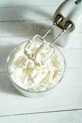 Whipped cream in a bowl and electric hand mixer on bright table.