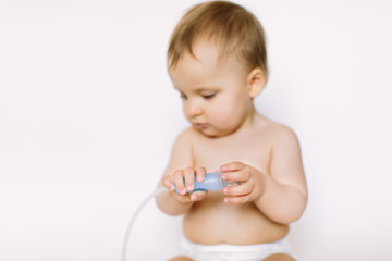 A little beautiful baby girl holds a nasal aspirator sitting on a white background. Hygiene and healthcare concept