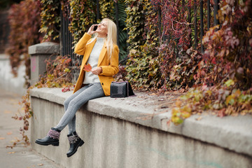 Young blond woman tourist is talikg on the mobile phone outdoor. Wearing bright yellow autumn coat, smiling. 