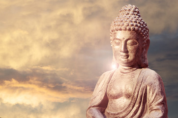Buddha statue sitting in meditation pose against sunset sky with golden tones fluffy clouds.