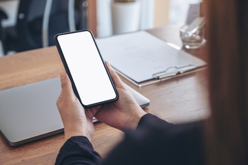 Mockup image of hands holding black mobile phone with blank white screen with laptop on wooden table in office
