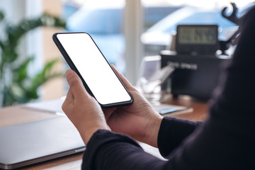 Mockup image of hands holding black mobile phone with blank white screen with laptop on wooden table in office