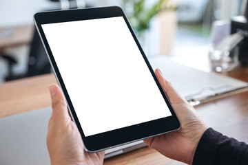 Mockup image of hands holding and using black tablet pc with blank white desktop screen with notebook on wooden table in office