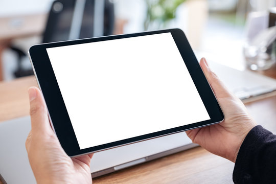 Mockup image of hands holding and using black tablet pc with blank white desktop screen with notebook on wooden table in office
