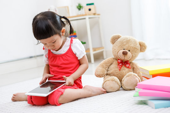 Preschooler girl plaing tablet. Cute child reading with teddy bear. Little girl having fun indoors at home, kindergarten or