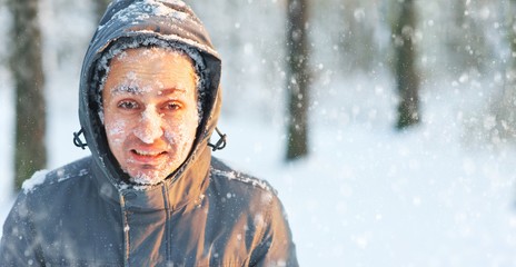 Frozen young man in a jacket with a hood covered with snow in winter forest. Snowflakes lie on the...