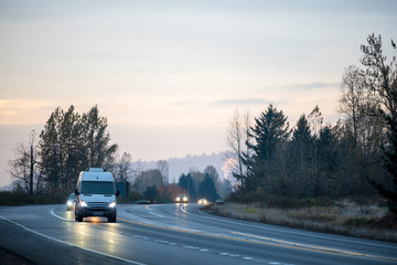Compact cargo mini van running on turning wet road in twilight with turned on headlight