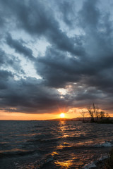 Obraz premium Beautufl sunset on a lake, with tree silhouettes and sun coming down under an overcast sky