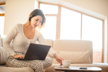 Pleasant blonde girl took a break in reading the article and reached for a cup of tea or coffee. The morning light fills the room with warm rays