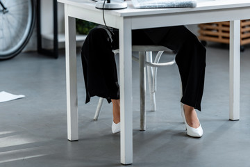 cropped view of woman in white shoes and trousers sitting at table at workplace