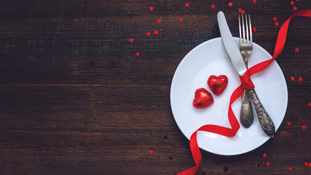 Valentine's Day Festive table setting, flat lay with two red heart shape chocolate candies on white plate, fork, knife and red ribbons on wooden table. Valentine Day, love, dating concept, copy space