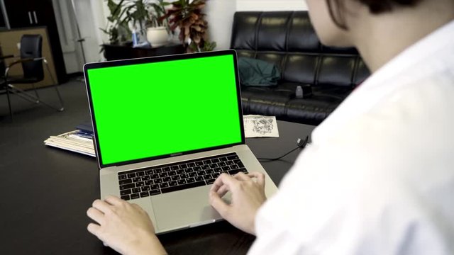 Over the shoulder shot of a woman using computer laptop with green screen display in office. Business woman using laptop with blank green screen on wooden table in office