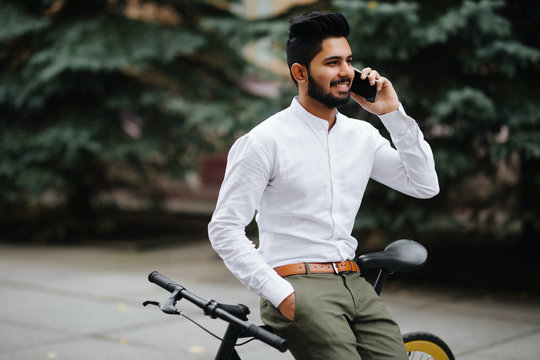 Handsome young indian man wearing suit talking on phone while walking with bicycle to work in the morning. Businessman going to work with his bicycle.