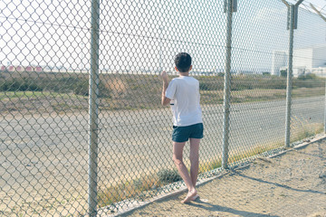 A barefoot boy stands on the beach near the iron net and looks at the airport.