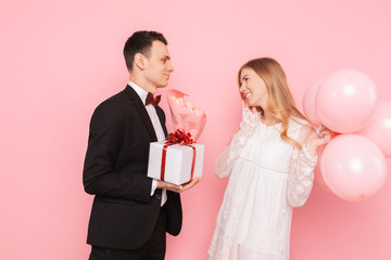 Elegant man in a suit, gives a box with a gift and a bouquet of flowers, to a beautiful woman, on a pink background. Women's day