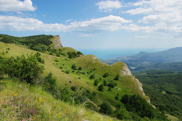 Fototapeta premium View from the Crimean Highlands on the seaside town and the Black Sea. Green cliff, forested mountain slopes and bright southern sky with light clouds