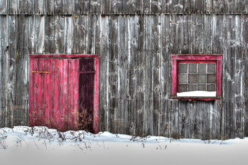Closeup of old gray wood planks barn in a snow field with red door and red window