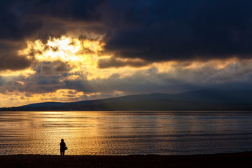 Naklejka na ściany i meble A lone figure in the sunset on the beach. Dramatic sunset, clouds, reflections in the sea, loneliness, thoughtfulness, reflections