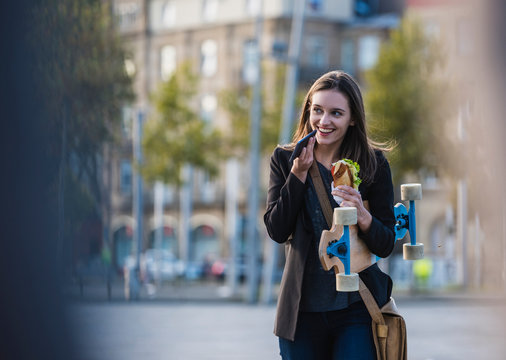 Smiling young woman with longboard and snack in the city using cell phone