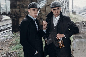 stylish gangsters men, smoking. posing on background of railway with bottle of alcohol.sherlock...