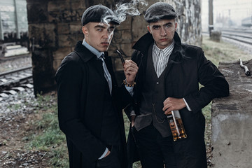 stylish gangsters men, smoking. posing on background of railway with bottle of alcohol.sherlock...