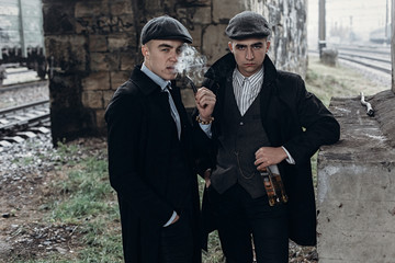 stylish gangsters men, smoking. posing on background of railway with bottle of alcohol.sherlock...