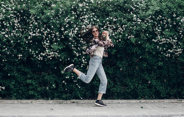 Fototapeta na wymiar happy hipster woman in sunglasses having fun on background of blooming bush with white flowers of spirea. boho girl jumping and smiling in modern clothes in summer street. space for text