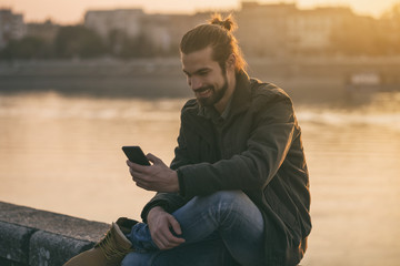 Handsome modern businessman using phone while sitting by the river.