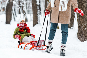 cropped view of mother pulling daughter in warm clothing on sledge in winter park