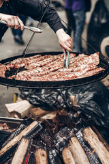 Fototapeta premium street food festival. grilled ribs meat. man holding steel tongues and roasting beef pork on big grill, under wooden fire. chef in black form cooking smoked juicy ribs. summer picnic