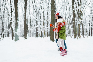 Fototapeta na wymiar attractive african american mother and cute daughter walking and holding cups in snowy forest