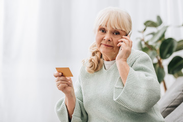 confused retired woman holding credit card while talking on smartphone