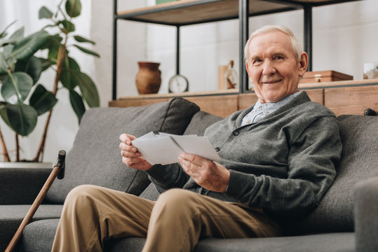 cheerful senior man with grey hair holding photos and sitting on sofa