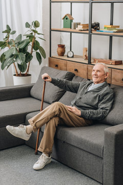 Happy Pensioner Smiling And Holding Walking Stick While Sitting On Sofa In Living Room