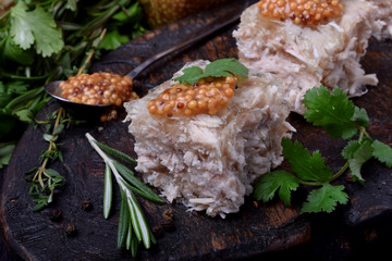 Holodets chicken jelly cut into cubes topped with mustard and cilantro on a dark wooden board surrounded by herbs and pepper