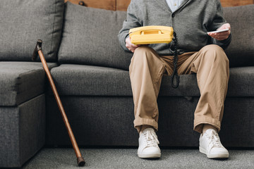 cropped view of retired man holding smartphone and retro phone while sitting on sofa