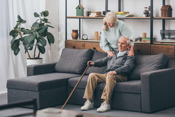 retired wife embrace sad senior husband sitting with walking cane in living room