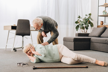 supportive retired husband looking at senior wife lying on floor and holding head