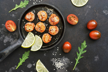 Grilled scallops on a cast-iron pan with lemon, arugula leaves and black cherry tomatoes, black stone background. Top view, flat lay.