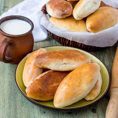 Homemade ruddy pies with filling on a round plate and in a wicker basket, next to a mug of milk.