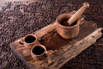 Mortar with ground coffee and pestle lie on a wooden board. Coffee pours from the mortar. Clay mugs...