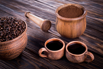 Two mugs of clay with coffee stand on a wooden board. Against the background of a mortar with ground coffee